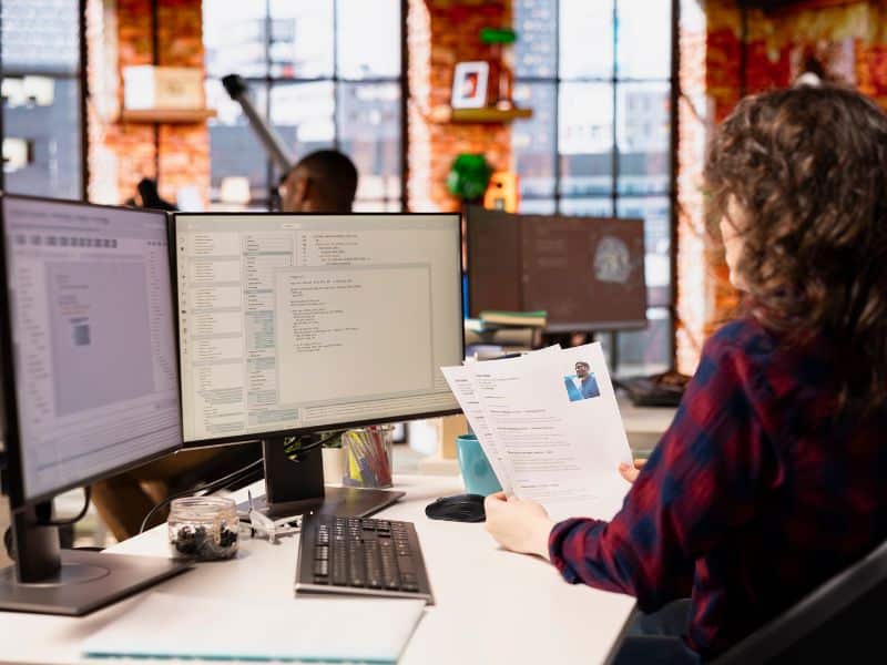A hiring manager sits behind multiple computer screens, holding a resume in her hands.