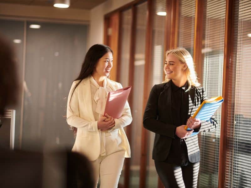 Two professionals walking and talking in an office hallway while holding work materials
